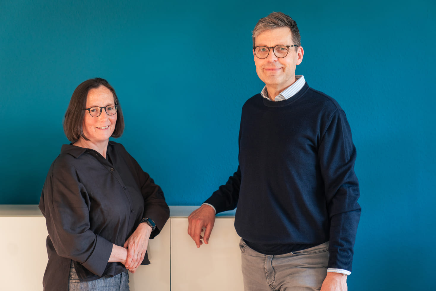 Dr. med. Annette Barwe und Dr. med. Georg Driesch vor einer petrolblauen Wand