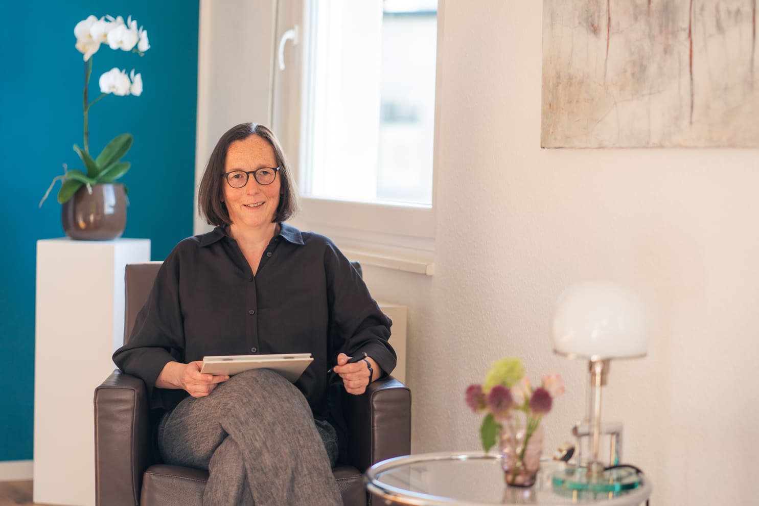 Dr. med. Annette Barwe sitzt mit einem Schreibblock im Ledersessel, hinter ihr eine weiße Orchidee vor petrolblauer Wand