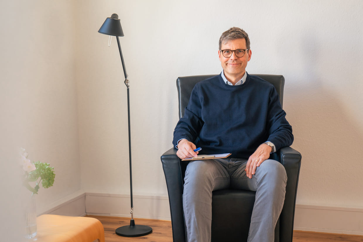 Dr. med. Georg Driesch sitzt mit einem Klemmbrett im Ledersessel neben einer Stehleuchte