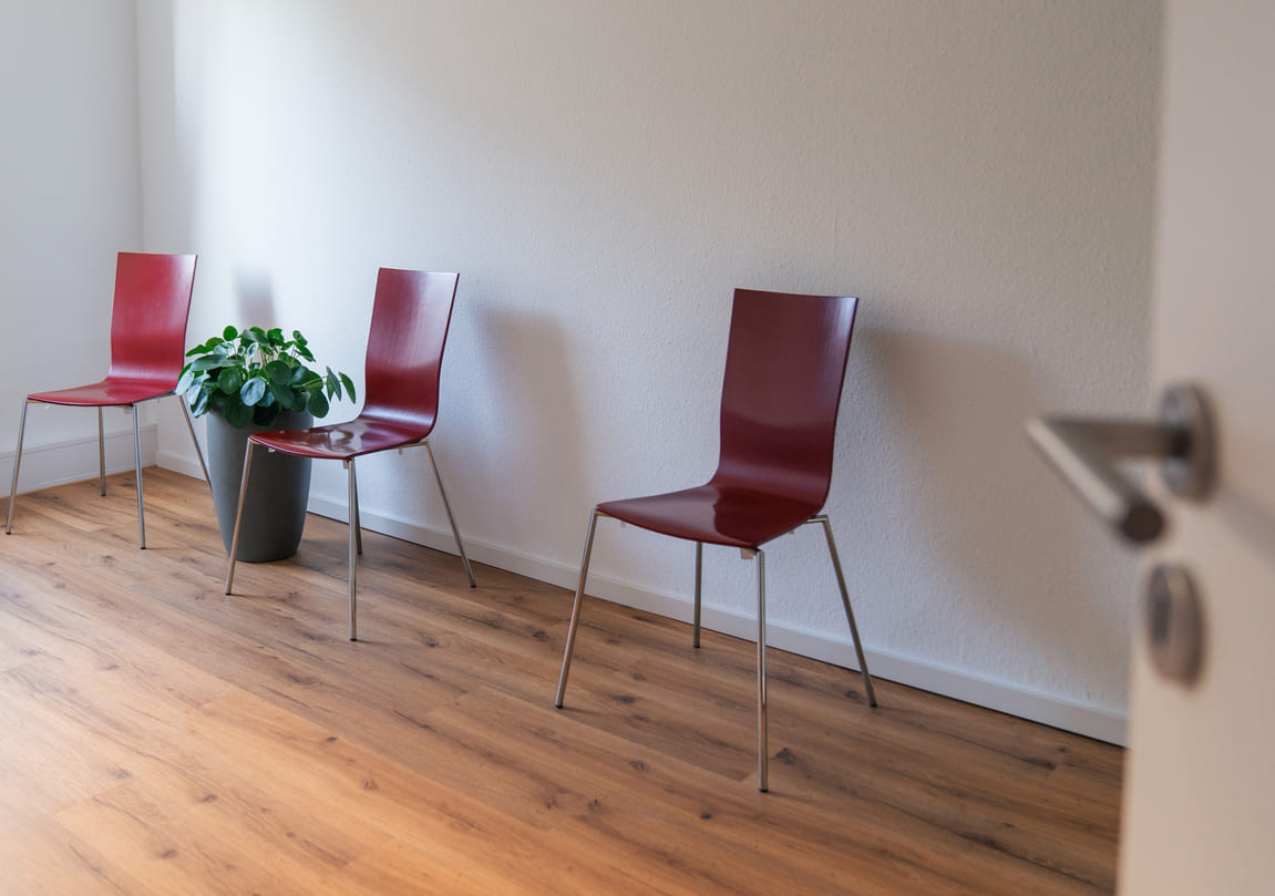 Wartezimmer mit drei roten Stühlen und einer Grünpflanze an einer weißen Wand